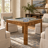 A wooden mahjong table with a blue felt playing surface and decorative tile borders, styled in a bright living room surrounded by beige armchairs and a patterned rug.