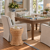 A styled living room scene featuring a wooden game table surrounded by light beige armchairs with dark piping, a small round wicker side table, and a woven rug. A board game is set on the table, and the room is lit by large windows with a view of water and shoreline.
