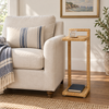 A tall, narrow woven side table with a natural rattan finish stands beside a cream upholstered armchair in a bright living room. The table holds a small glass and a book on its lower shelf.