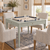 A light sage green folding game table surrounded by four striped armchairs in a bright, coastal-style living room. The table is set up with a game board and pieces, with glasses and small bowls on top.