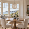 A round wooden dining table with four upholstered beige chairs in a bright coastal dining room, featuring a shell-like chandelier, bay windows with a water view, and floral centerpieces.