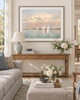 A framed horizontal canvas print of a sailboat sunset scene, displayed on a white wall above a wooden console table in a bright, modern living room.