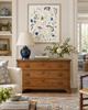 Framed blue, green and blush botanical artwork displayed above an antique wood chest in a traditional light-filled living room.